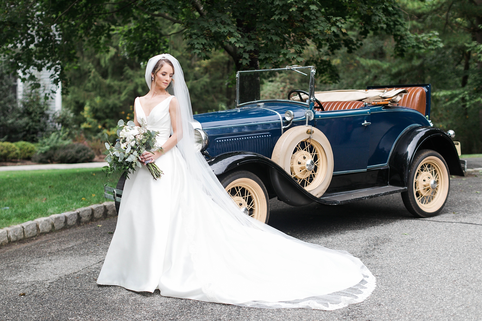 bride poses in front of blue antique car with long train and veil and green and white wedding florals