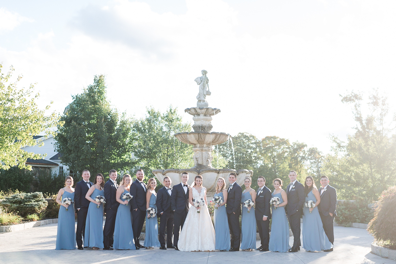 bridal party on wedding day with blue dresses and tuxedos pose with bride and groom in front of fountain at Trump National Golf Club Westchester
